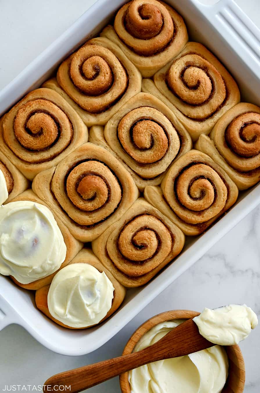 A top-down view of easy Make-Ahead Cinnamon Rolls with Cream Cheese Frosting in a white baking dish next to a bowl of the best cinnamon roll frosting