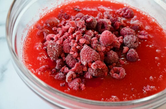 A glass bowl containing raspberry Jell-O and frozen raspberries