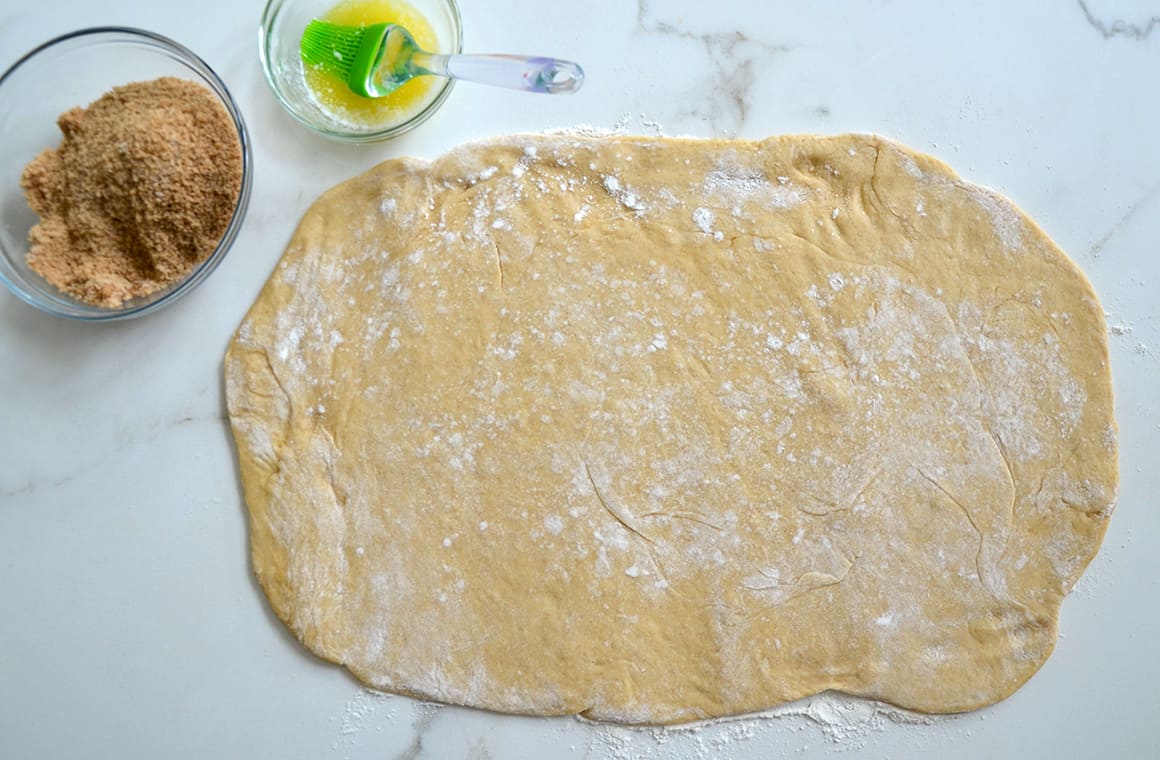 A top-down view of easy cinnamon roll dough next to small bowls containing melted butter and a cinnamon-sugar mixture