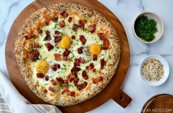 Top down view of Everything Bagel Breakfast Pizza on wood board next to dish towel and small ramekins containing chives and everything seasoning