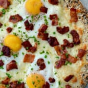 A top-down view of Everything Bagel Breakfast Pizza topped with crispy bacon and three fried eggs.