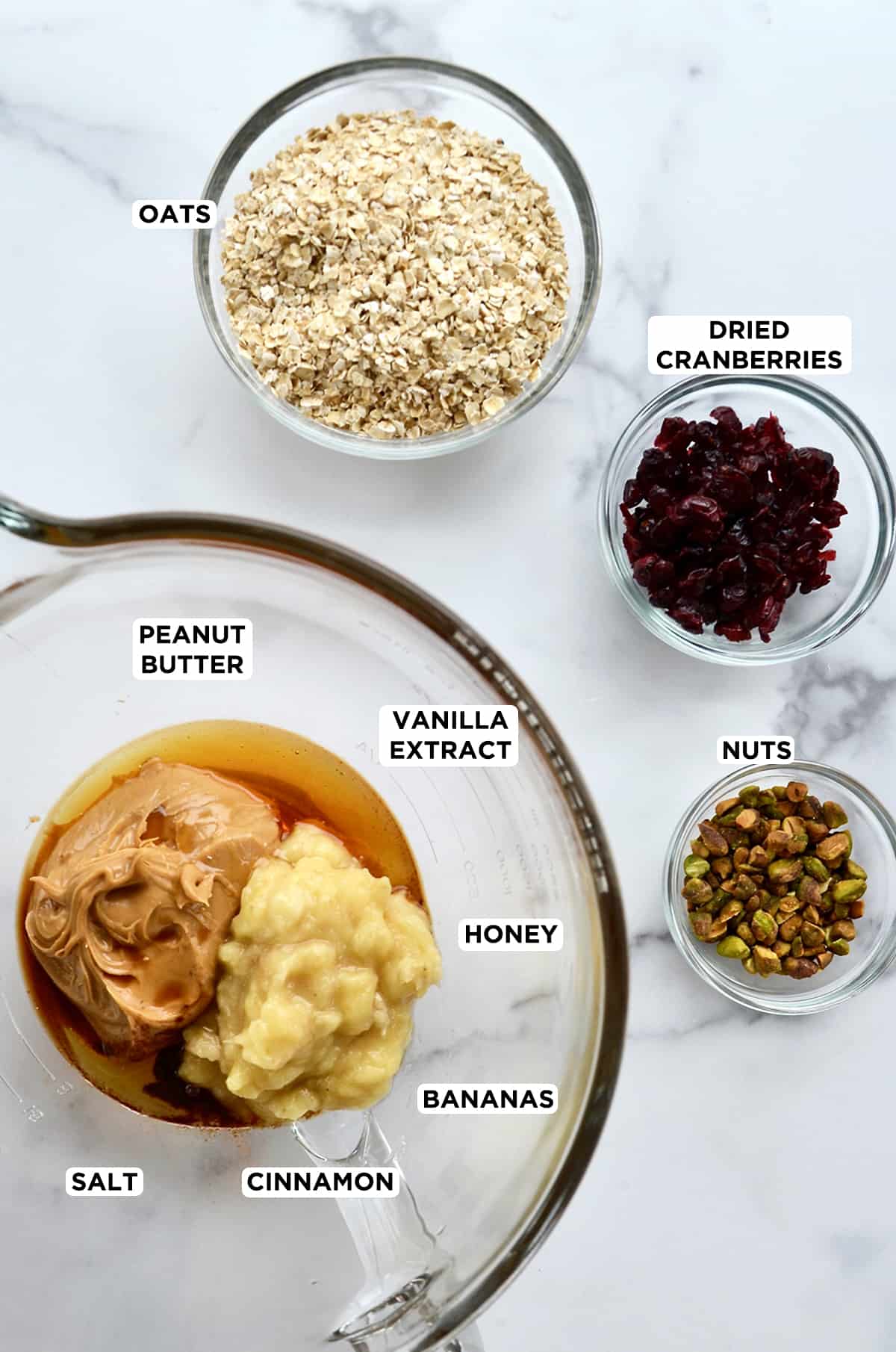 Ingredients for breakfast cookies in glass bowls, including quick oats, dried cranberries, chopped nuts, creamy peanut butter, mashed bananas, vanilla extract and honey.