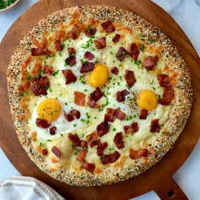 Everything Bagel Breakfast Pizza on wood board next to pizza cutter, dish towel and small ramekins containing herbs and spices