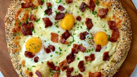 Everything Bagel Breakfast Pizza on wood board next to pizza cutter, dish towel and small ramekins containing herbs and spices