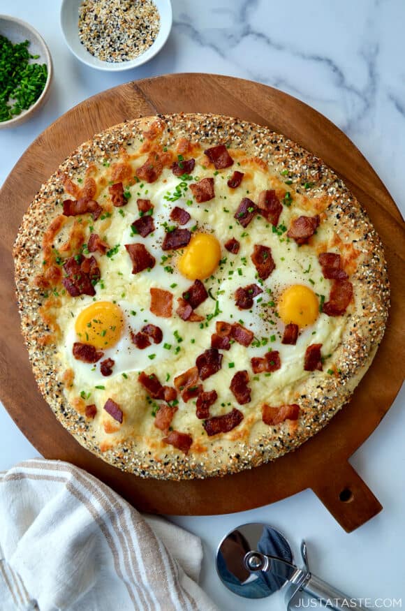 Everything Bagel Breakfast Pizza on wood board next to pizza cutter, dish towel and small ramekins containing herbs and spices