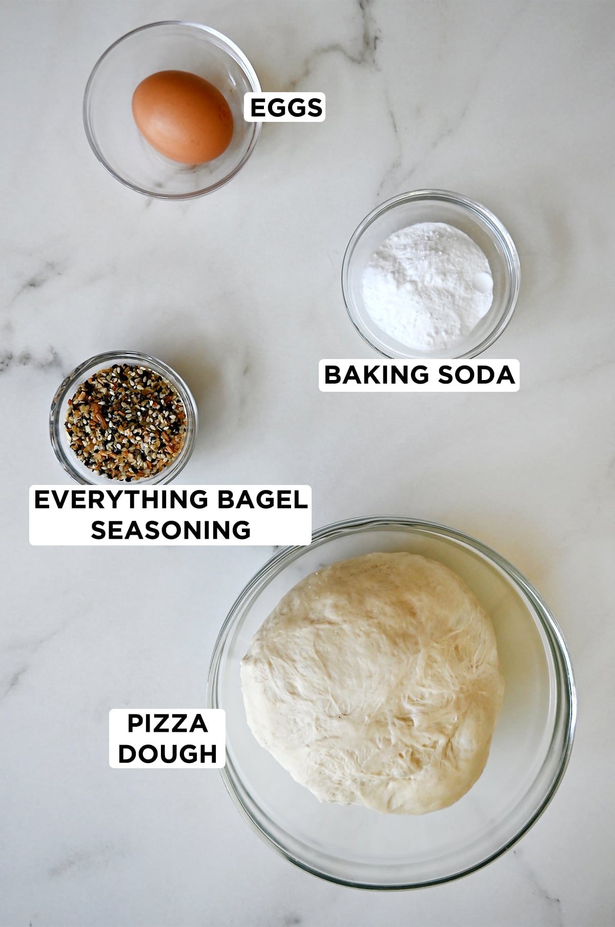Various sizes of clear bowls containing an egg, baking soda, pizza dough and everything bagel seasoning.