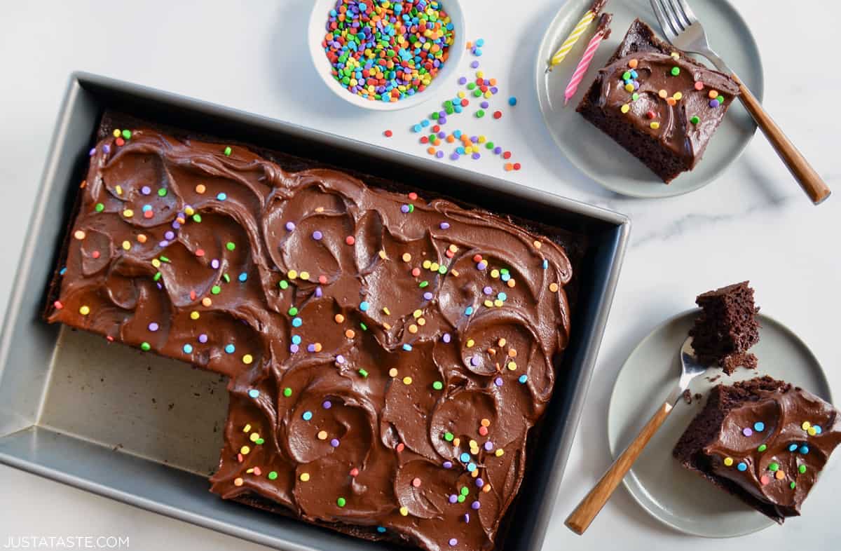 A chocolate sheet cake topped with silky chocolate buttercream and bright rainbow sprinkles with two pieces of cake on plates next to it.