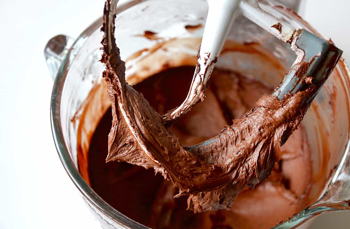 Creamy chocolate buttercream covering a paddle attachment of a stand mixer with more buttercream in the bowl.