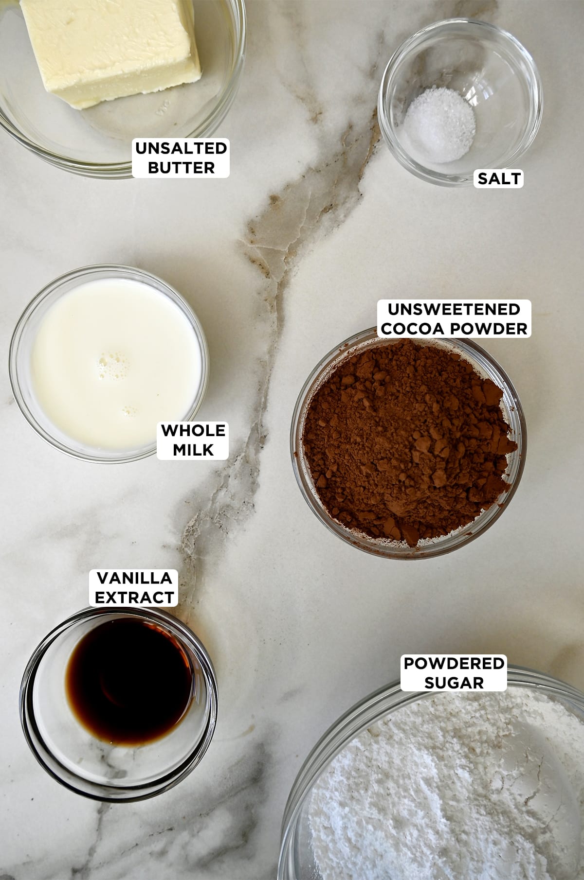 Glass bowls containing ingredients to make chocolate buttercream frosting, including room temperature butter, salt, unsweetened cocoa powder, powdered sugar, vanilla extract and whole milk.