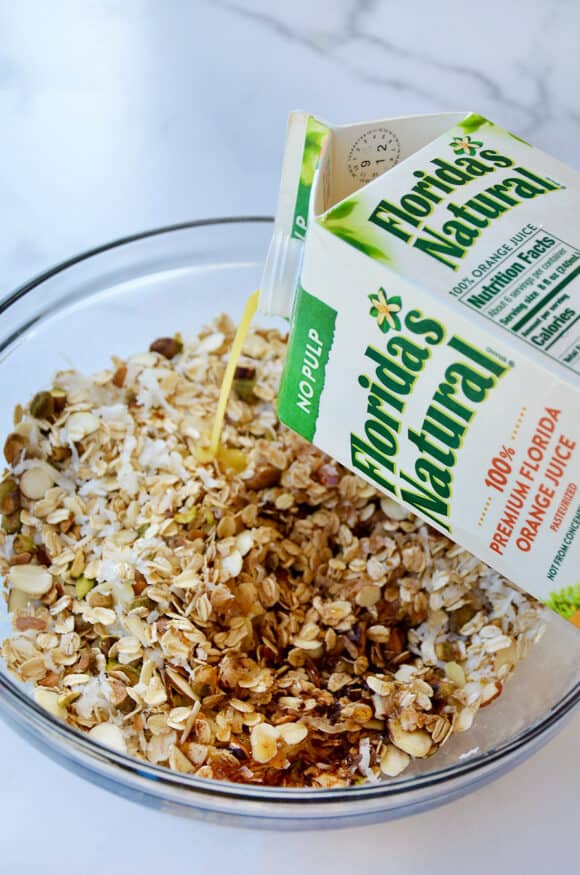 A carton of orange juice being poured into a bowl with granola ingredients