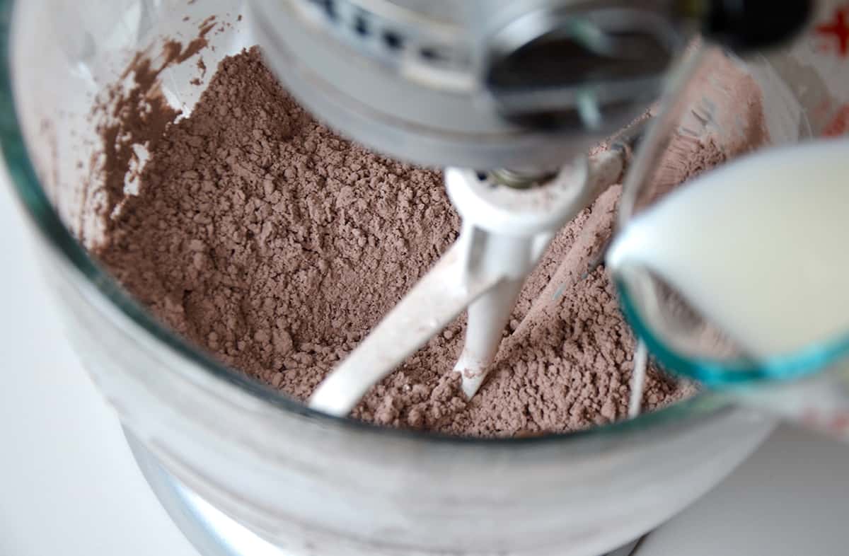 Milk being streamed into a stand mixer bowl that contains cocoa powder, powdered sugar and butter.