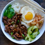 Easy Beef Bulgogi in a bowl with white rice topped with a fried egg, pickled cucumber, kimchi and fresh cilantro