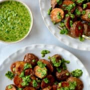 A top-down view of two white plates containing roasted potatoes topped with chimichurri sauce