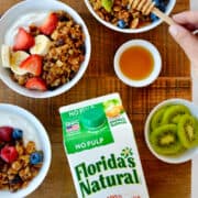 A top-down view of a carton of Florida's Natural Orange Juice next to granola parfaits with fruit and yogurt next to small bowls containing blueberries, honey and slices of kiwi.