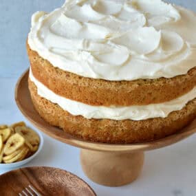 A two layer banana cake with a thick layer of cream cheese frosting in the middle and on the top on a wooden cake stand.