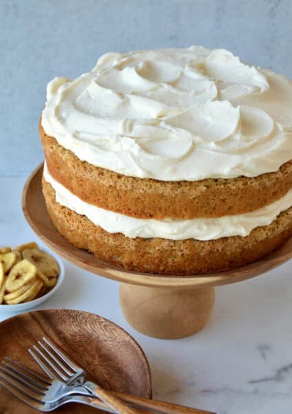 A two layer banana cake with a thick layer of cream cheese frosting in the middle and on the top on a wooden cake stand.