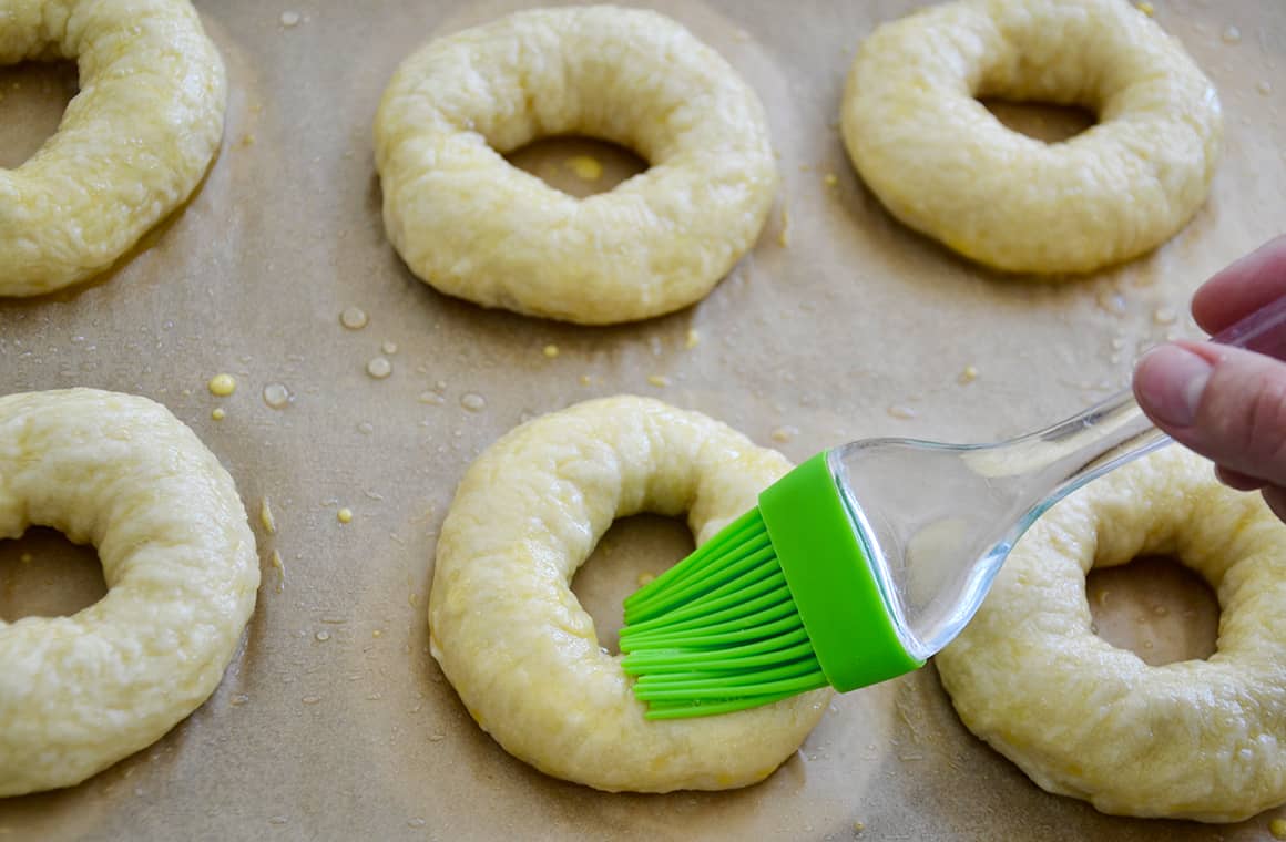 Soft pretzel bagels on a baking sheet being brushed with egg wash