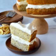 A slice of banana cake topped with cream cheese frosting on a plate with the rest of the cake on a cake stand in the background.