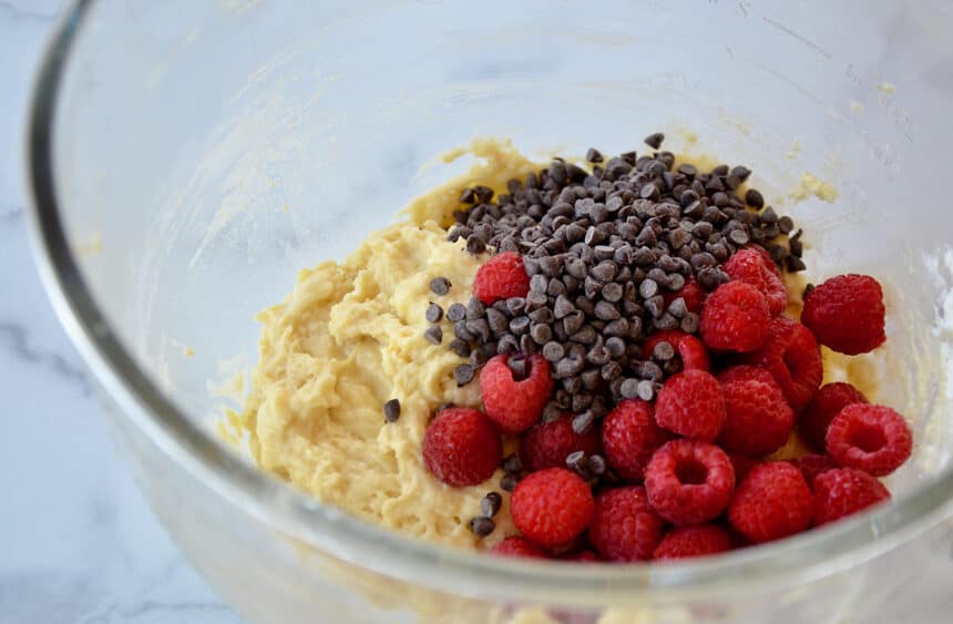 A glass bowl containing muffin batter, raspberries and mini chocolate chips