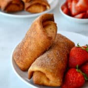 A close-up view of two Strawberry Cheesecake Chimichangas on a white plate with fresh strawberries.