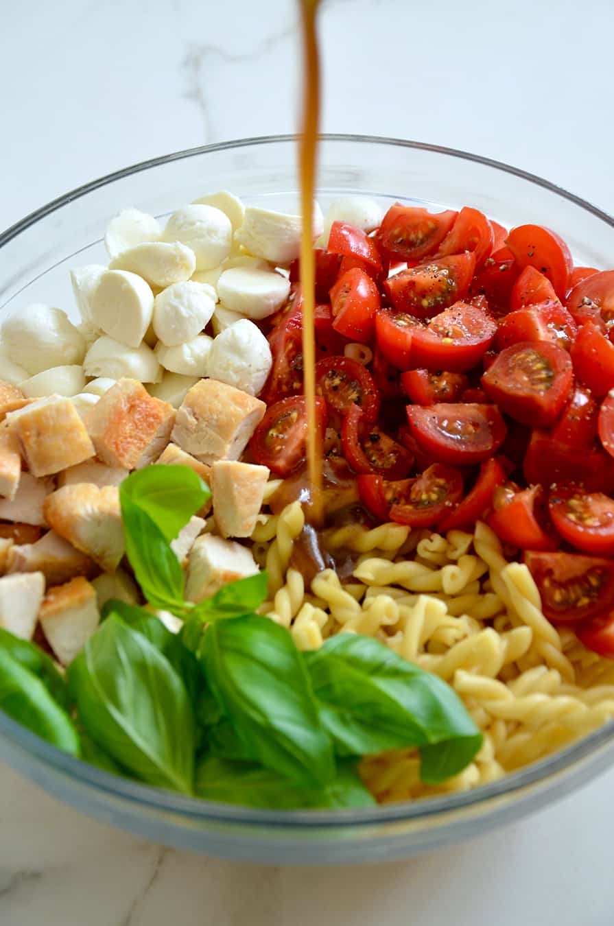 A glass bowl containing pasta, basil, mozzarella and tomatoes with dressing being poured in