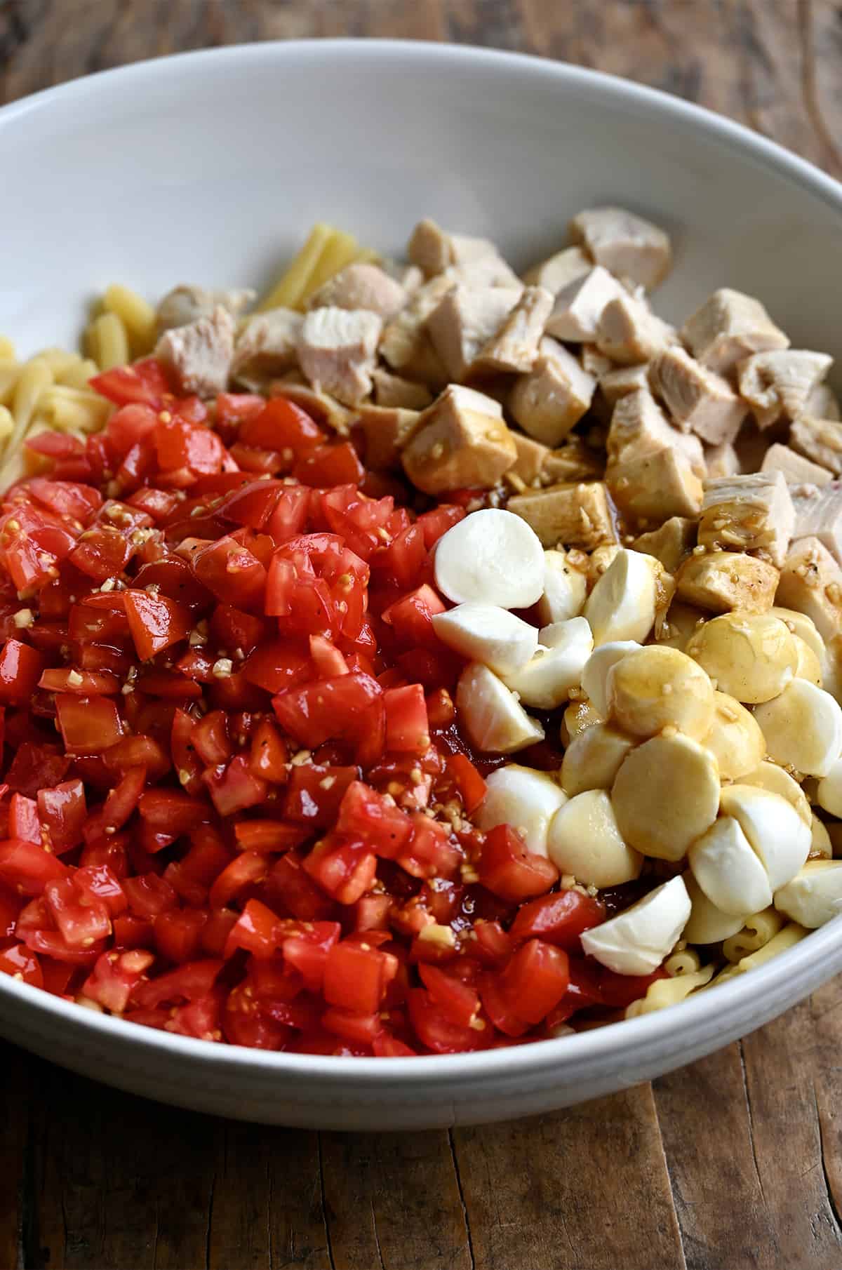 A large serving bowl containing diced tomatoes, cubed chicken, halved mini mozzarella balls and cooked noodles.