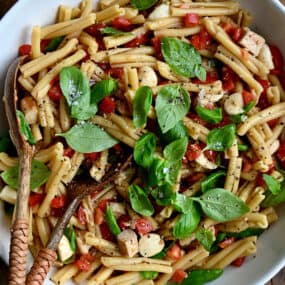 A top-down view of a large serving bowl containing Caprese pasta salad garnished with fresh basil leaves.