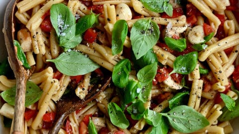 A top-down view of a large serving bowl containing Caprese pasta salad garnished with fresh basil leaves.