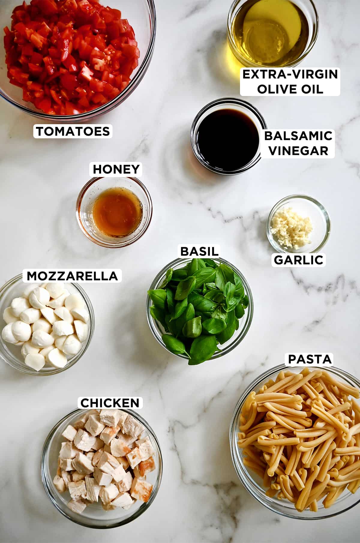 A top-down view of various sizes of clear bowls containing ingredients for Caprese pasta salad, including tomatoes, olive oil, balsamic vinegar, garlic, basil, honey, pasta, chicken and mini mozzarella balls.
