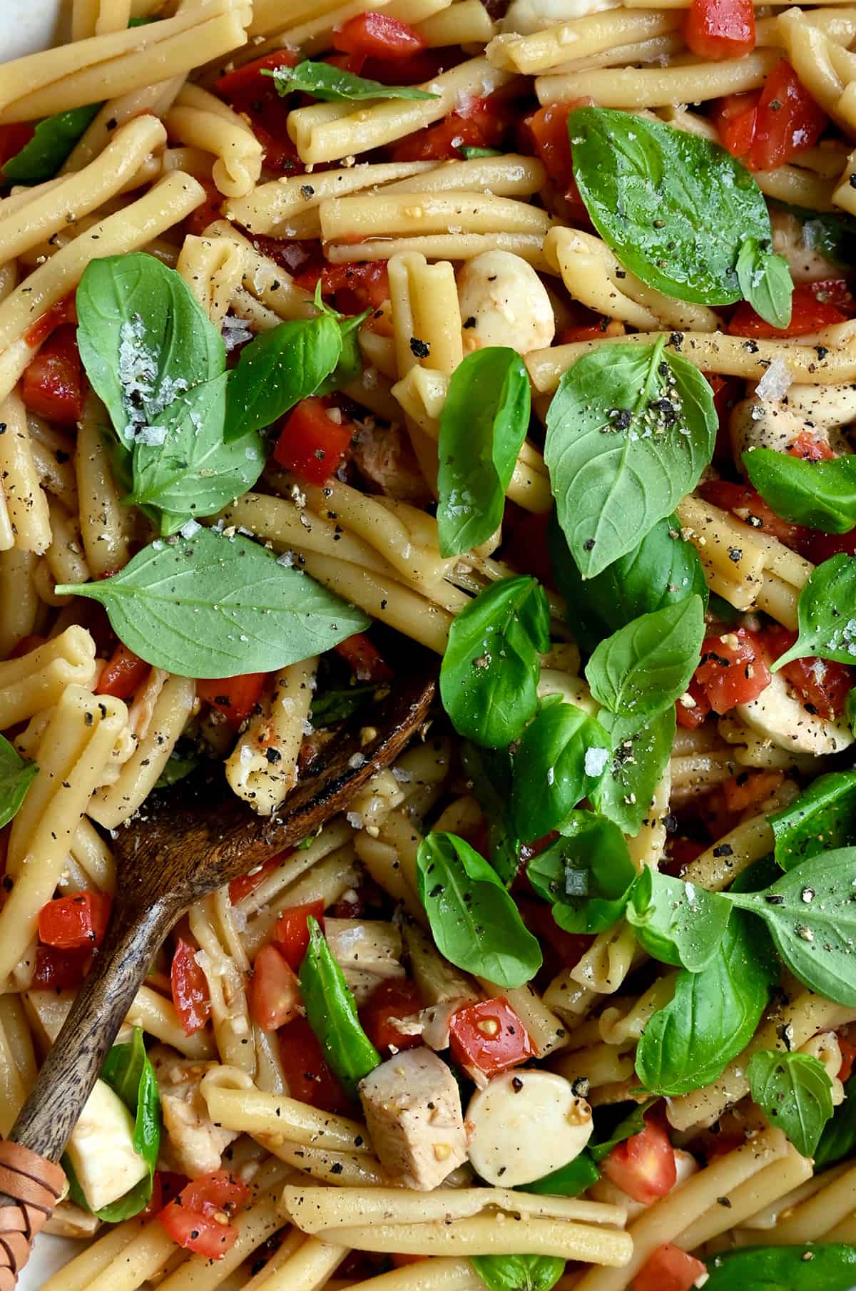 A close-up view of pasta salad with fresh basil, diced tomatoes, mini mozzarella balls and cubed chicken tossed in a garlicky balsamic dressing.