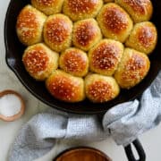 Dishcloth wrapped around the handle of a cast-iron skillet containing pull-apart pretzel rolls next to a small bowl filled with sea salt.