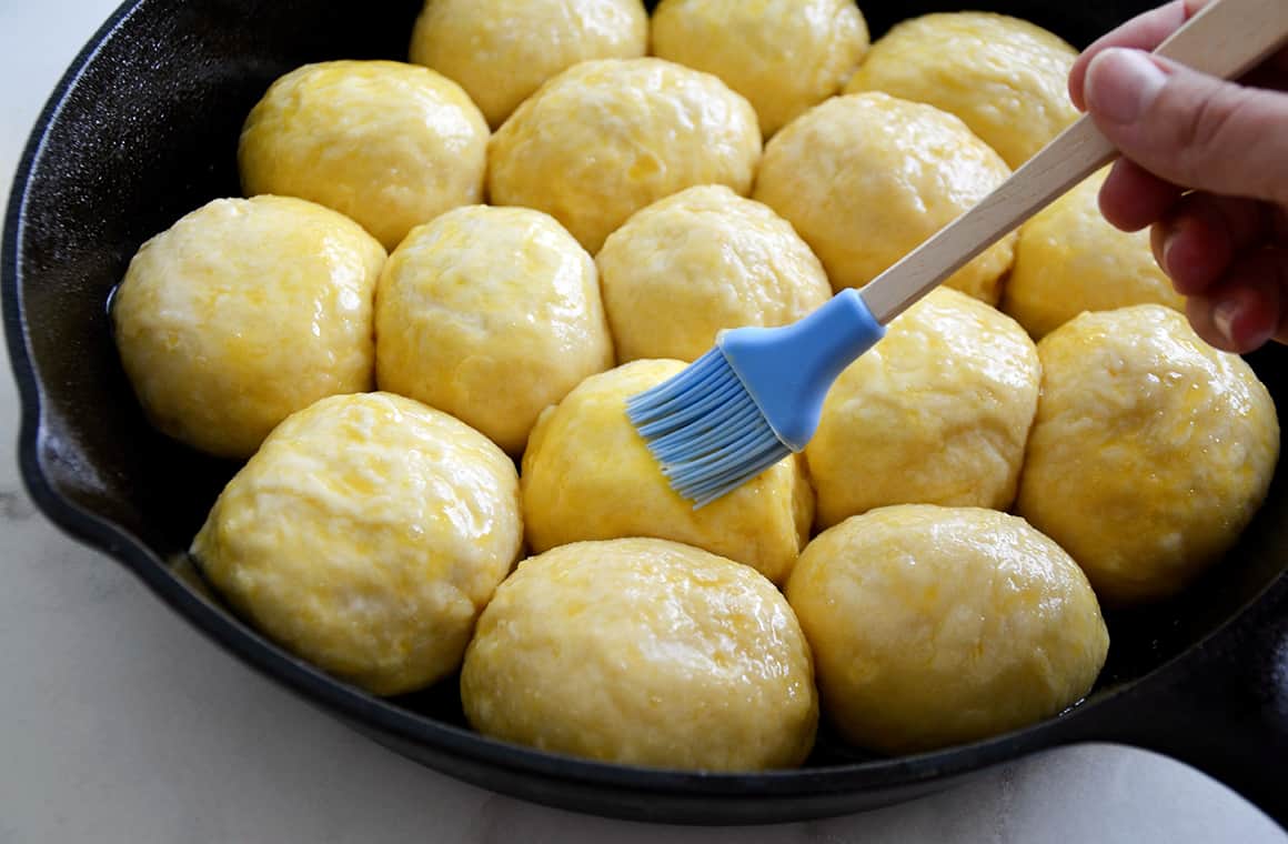 Pretzel roll dough balls nestled in a cast-iron skillet being brushed with egg wash