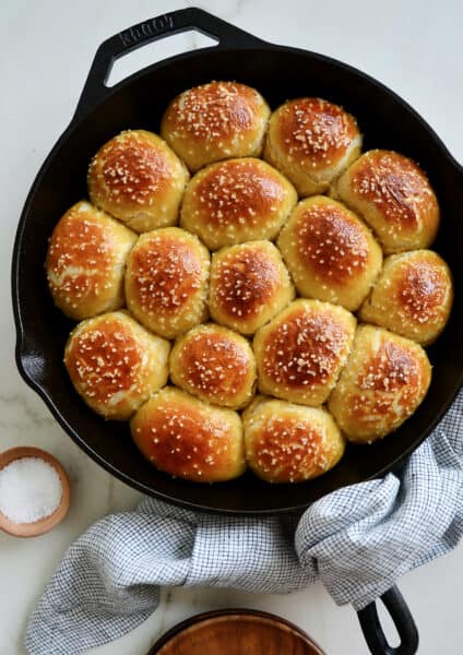 A cast-iron skillet filled with pretzel rolls with a small bowl of salt