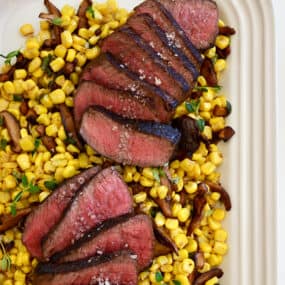 A white platter with corn, mushrooms and sliced steak