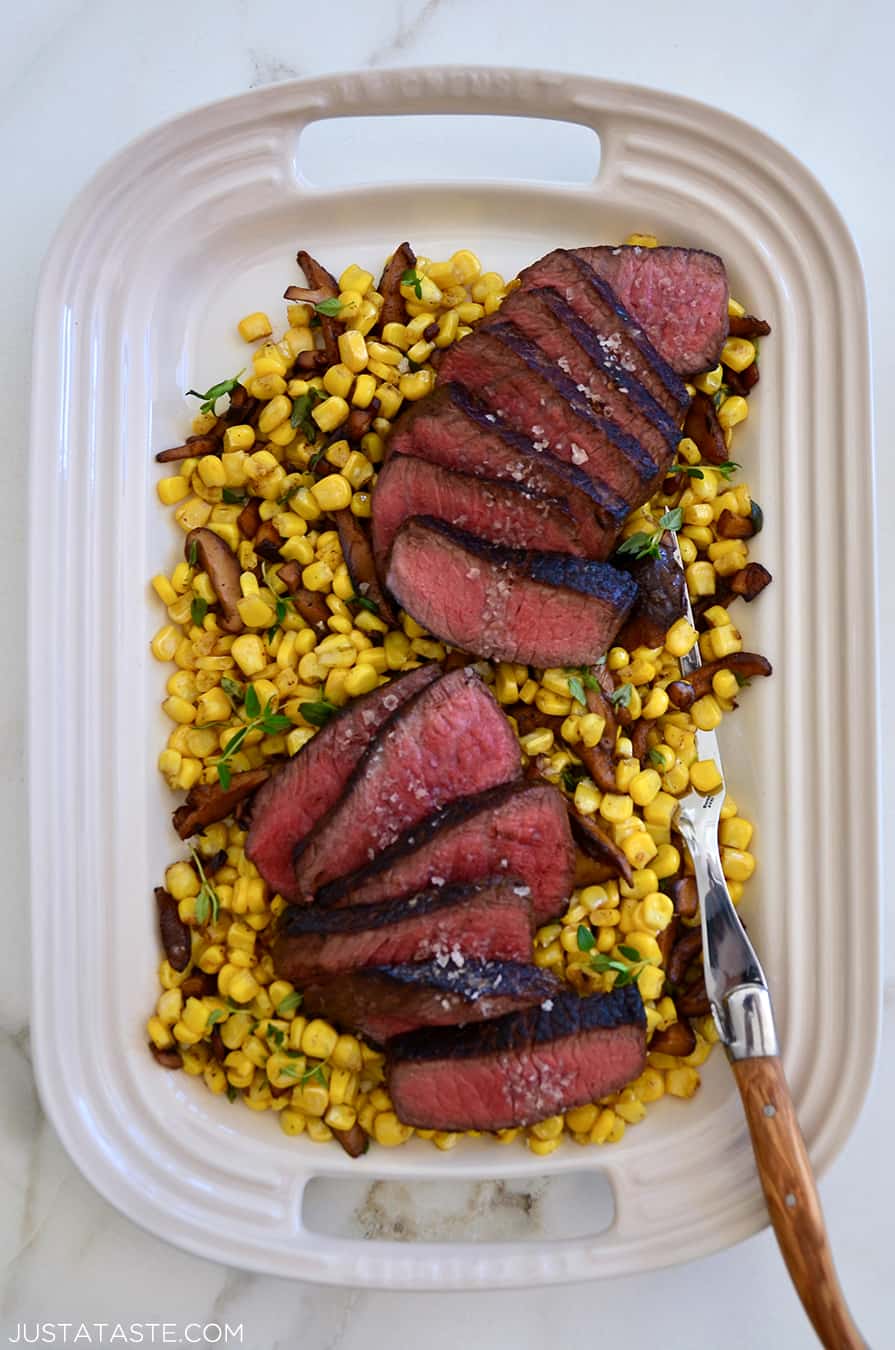 A white platter with sliced steak on top of corn and mushrooms