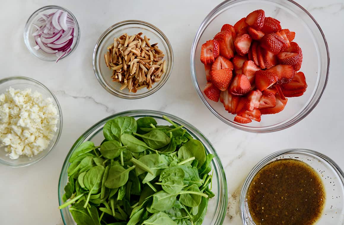 Clear glass bowls containing spinach, strawberries, onions, nuts and feta