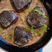 Four cooked steaks with fresh thyme in a skillet.