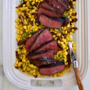A top-down view of sliced steak garnished with large-flake sea salt over a bed of roasted corn.