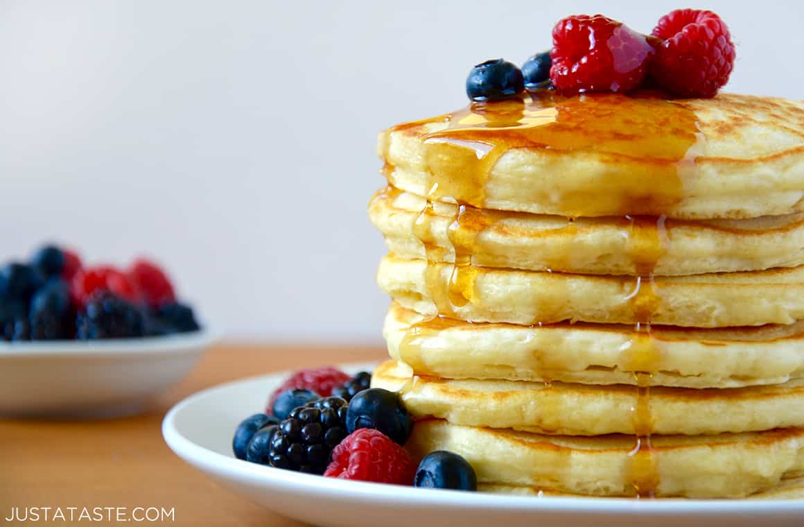A stack of Fluffy Greek Yogurt Pancakes on a white plate with fruit and syrup