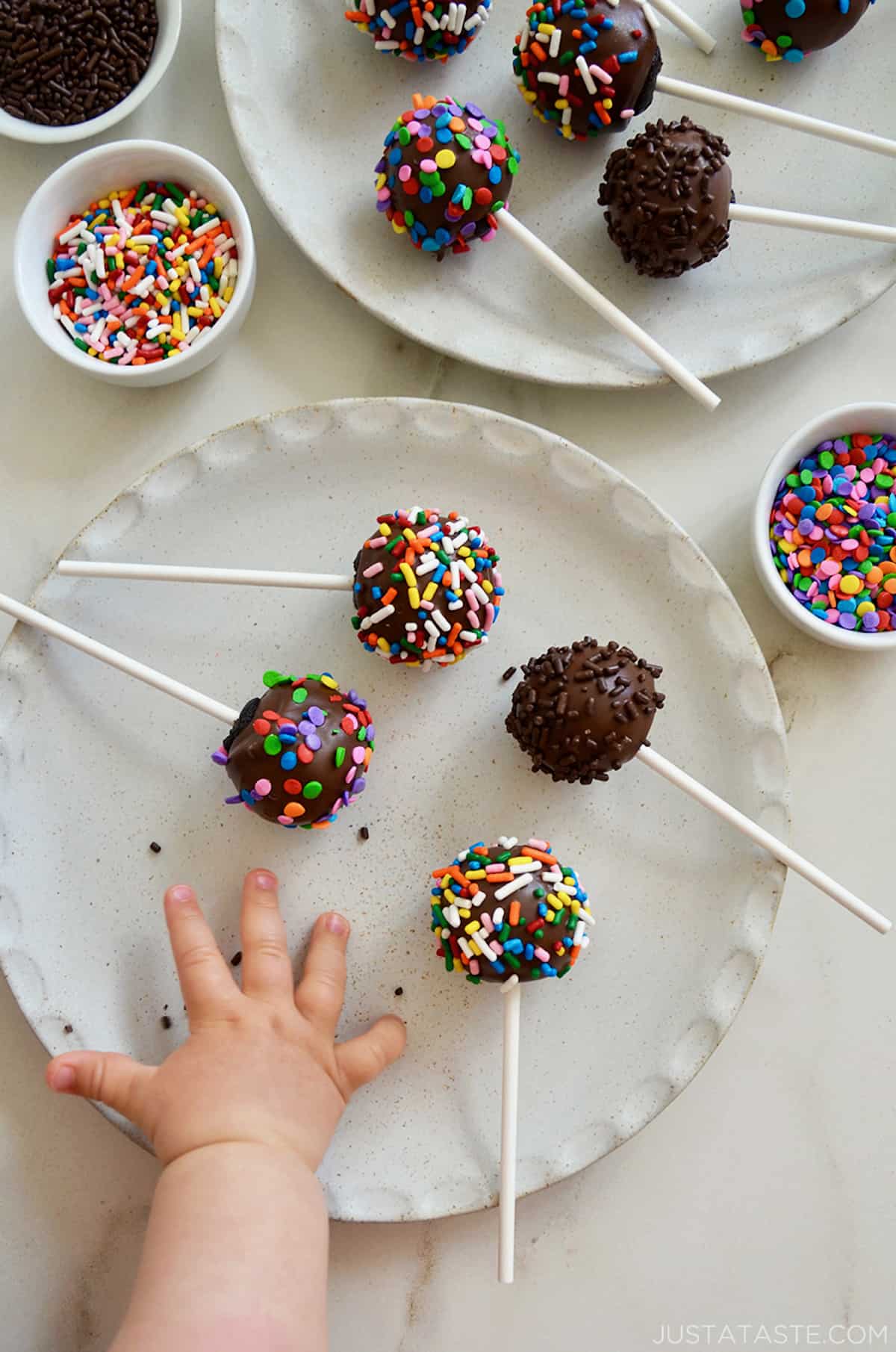 A child's hand reaches for a chocolate cookie pop with sprinkles on a white plate.