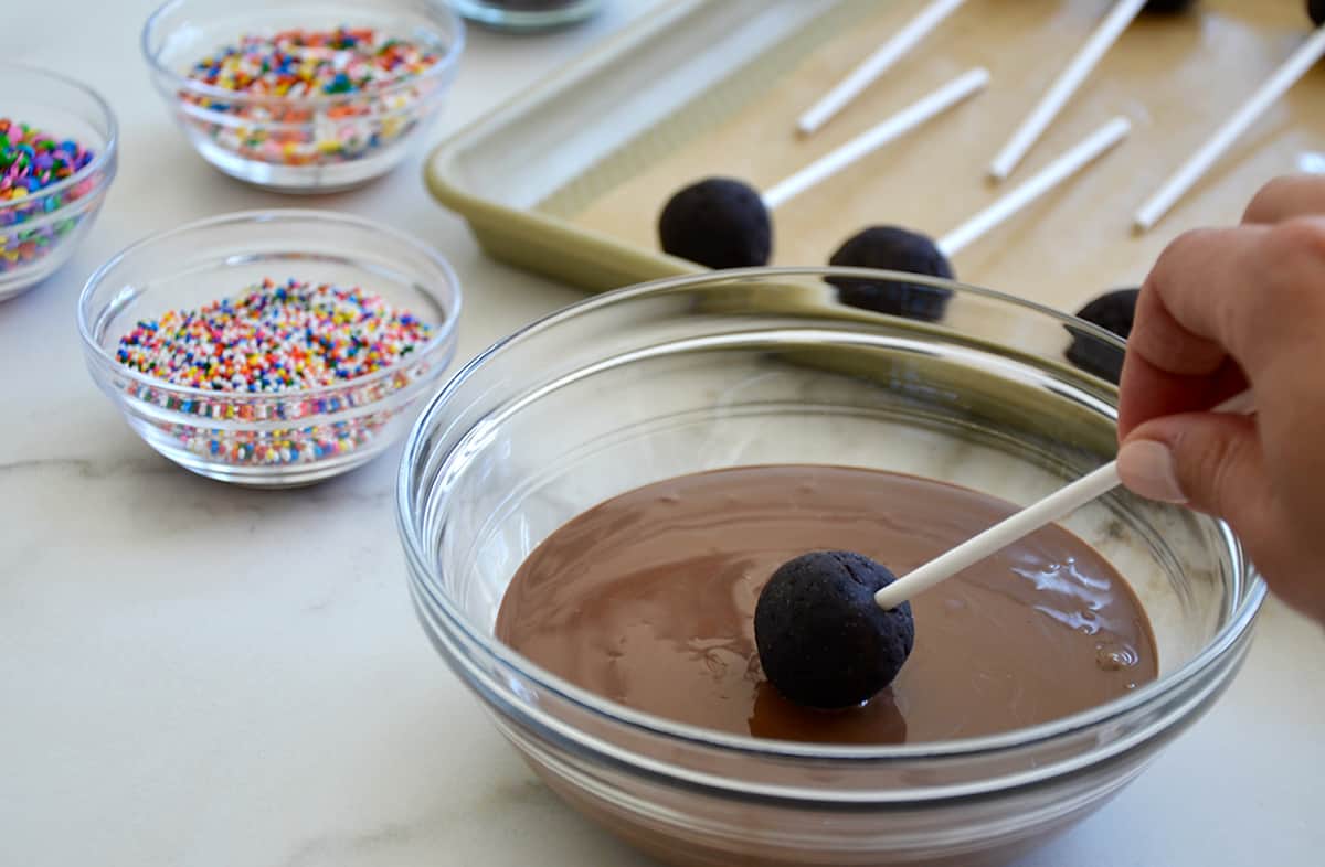 A chocolate cookie pop being dipped into melted chocolate in a clear bowl.