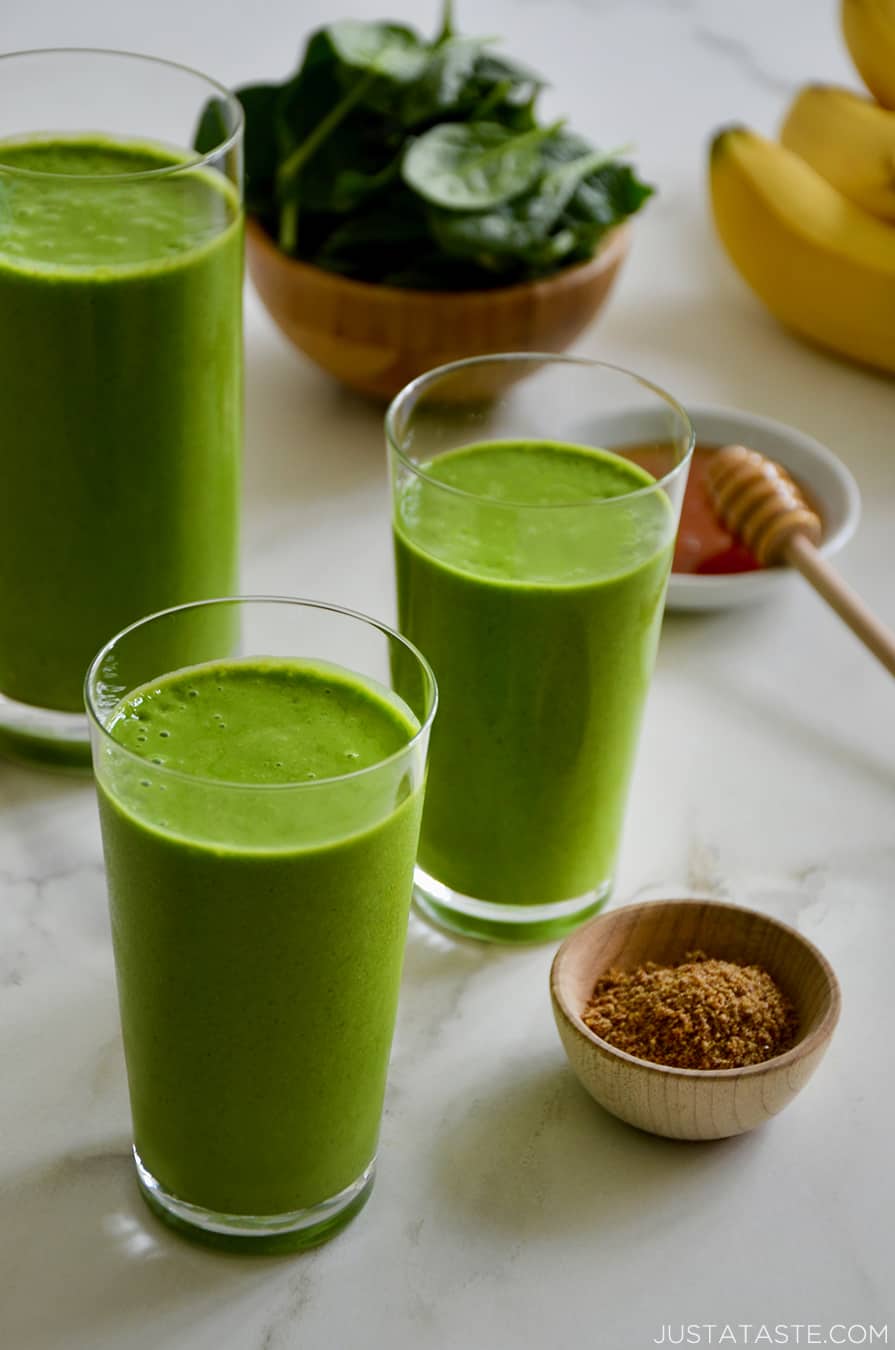 Three glasses filled with Healthy Green Smoothies next to a bowl containing fresh spinach, a small bowl filled with honey and another small bowl containing ground flaxseed.