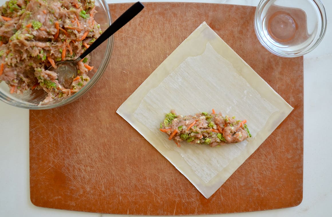 Ground chicken mixture atop a single egg roll wrapper next to a bowl containing a spoon and ground chicken mixture