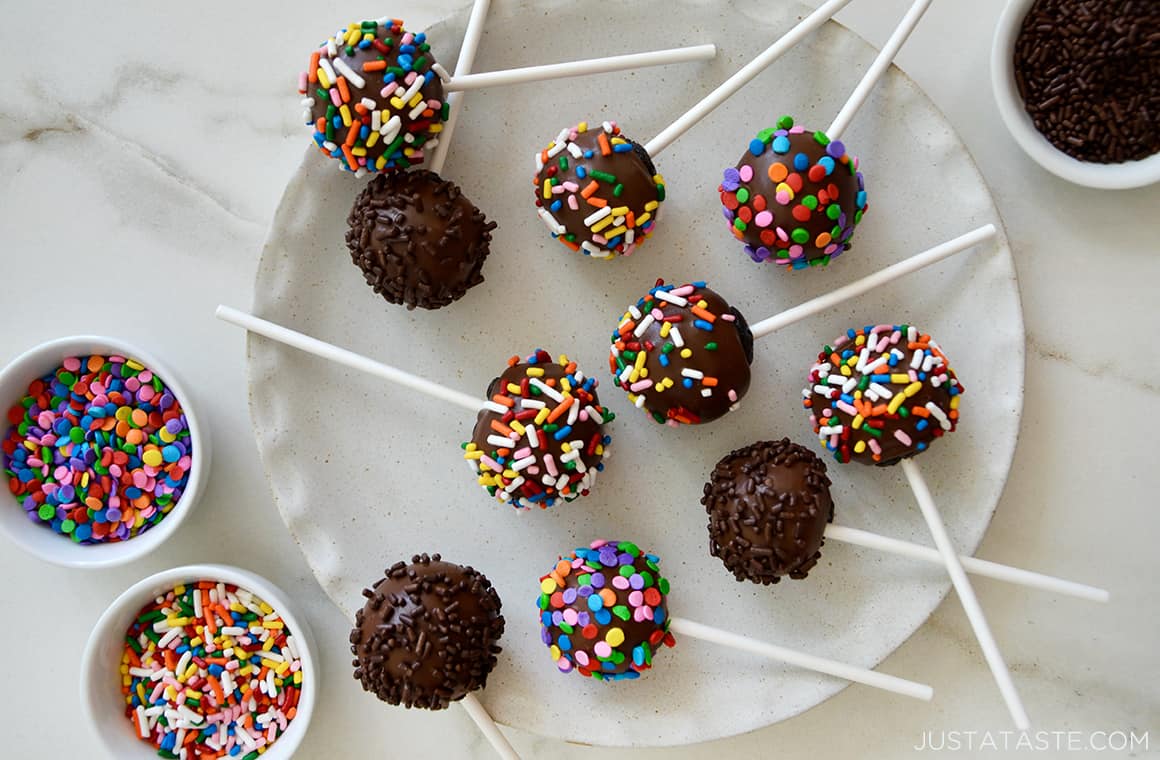 Chocolate cookie pops with sprinkles on a white dinner plate next to two small bowls containing sprinkles.