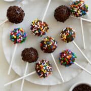 No-Bake Chocolate Cookie Pops decorated with rainbow and chocolate sprinkles on a white plate next to small bowls containing rainbow sprinkles.