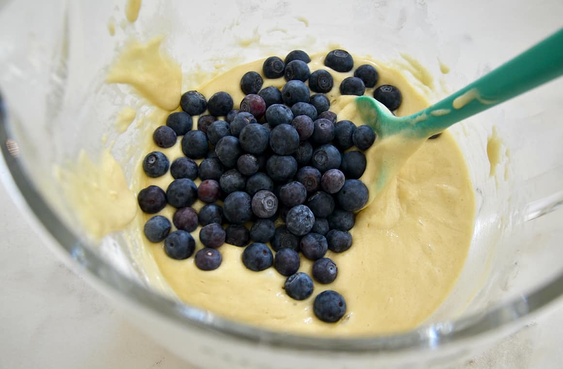 A glass bowl containing bread batter, blueberries and a spatula