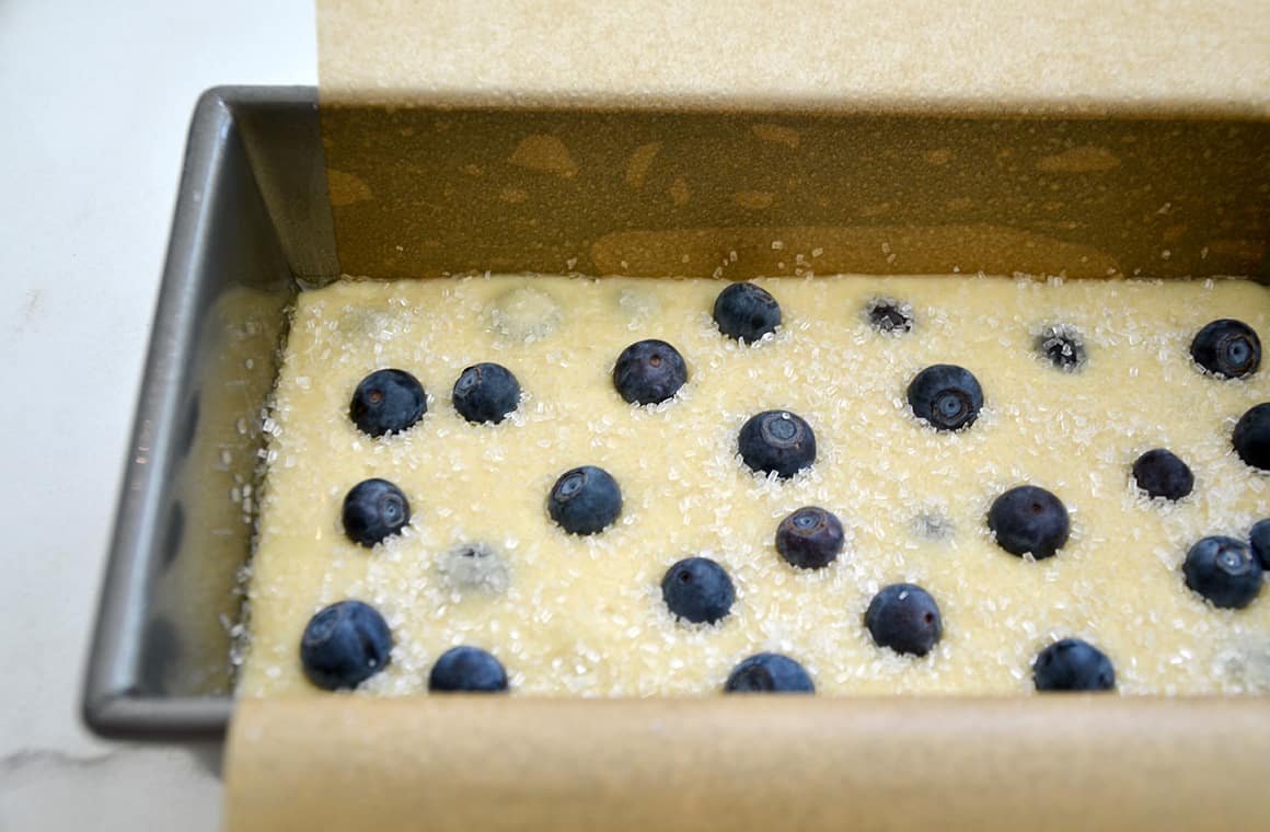 A loaf pan lined with parchment paper containing bread batter, blueberries and sanding sugar