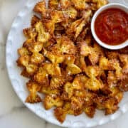 A top-down view of a white serving plate piled high with Parmesan Pasta Chips next to a small ramekin containing marinara sauce.