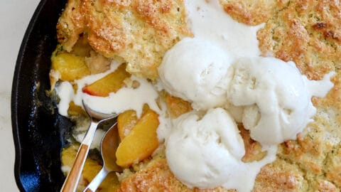 A skillet with peach cobbler and melting ice cream on top with spoons