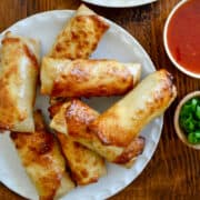 A white plate piled high with Air Fryer Egg Rolls next to small bowls containing sweet chili sauce and sliced scallions, and a plate with green lettuce topped with two egg rolls.